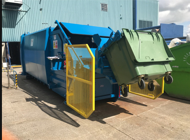 Industrial waste compactor lifting a large green wheelie bin with automated hydraulic arms outside a blue metal-clad building. The compactor is equipped with yellow safety guards and designed for high-volume waste processing.